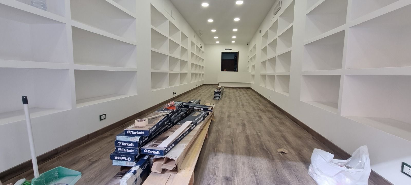Una stanza lunga e stretta con scaffali bianchi incorporati su entrambi i lati; materiali da costruzione giacciono sul pavimento.