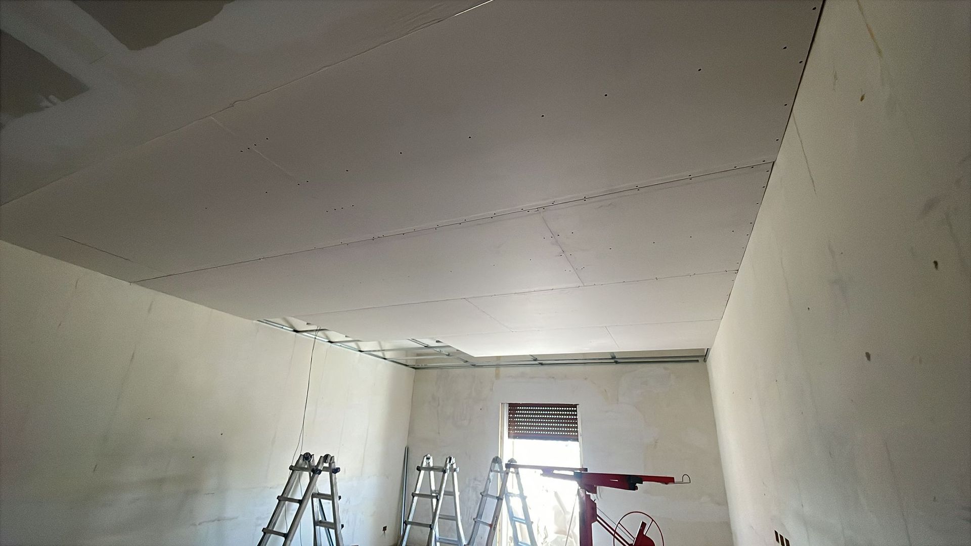 Una stanza con cartongesso sul soffitto e sulle pareti; sono presenti scale e un sollevatore per cartongesso.