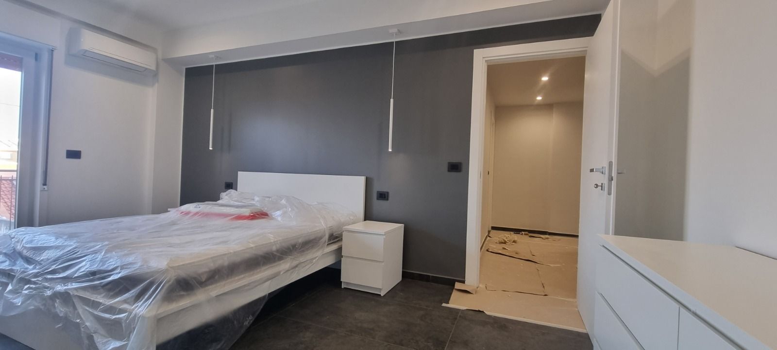 Camera da letto con parete grigia, letto bianco, comodino e porta aperta su un'altra stanza.