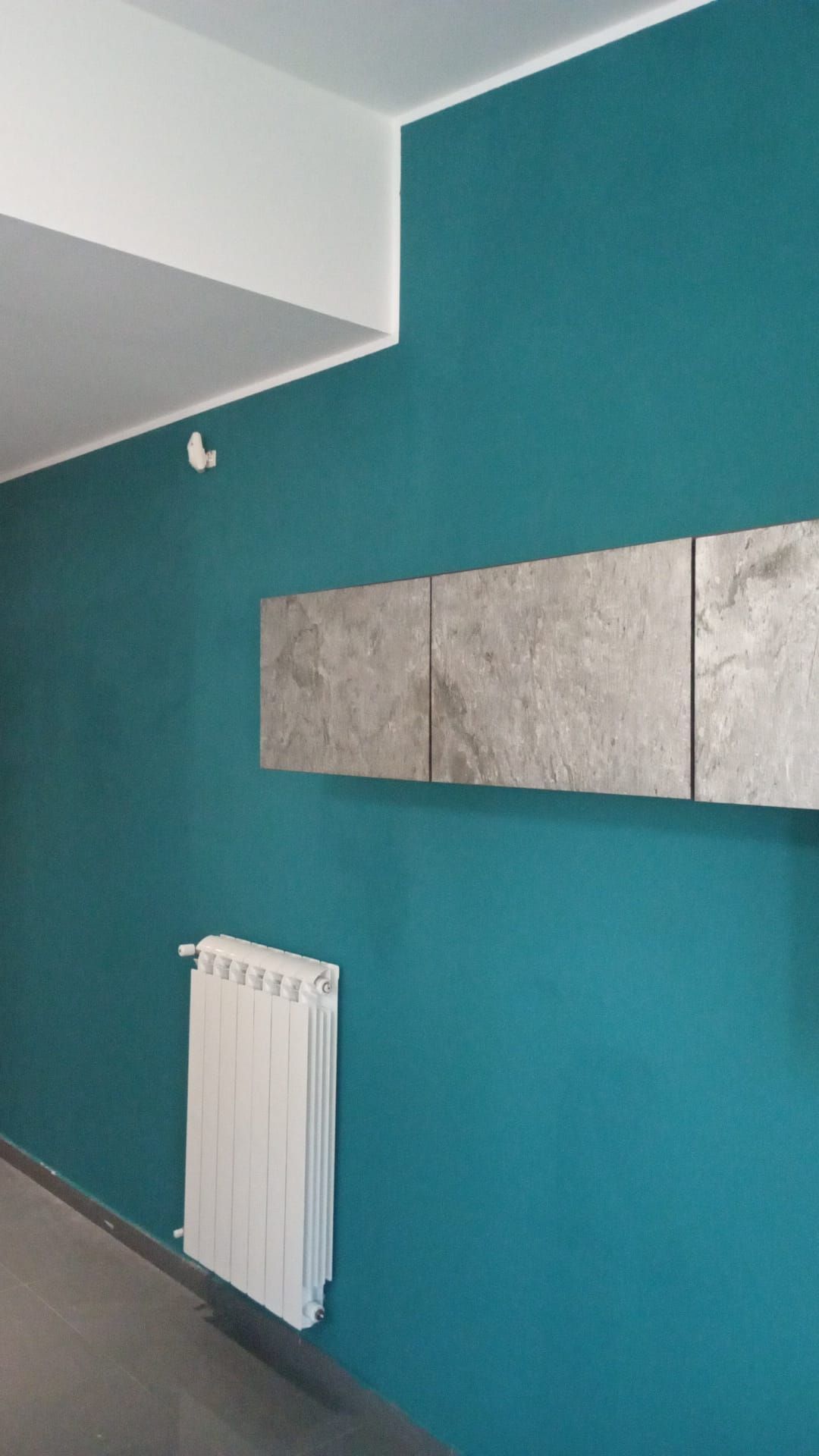 Parete color verde acqua con decorazione murale rettangolare grigia, termosifone bianco e soffitto bianco.