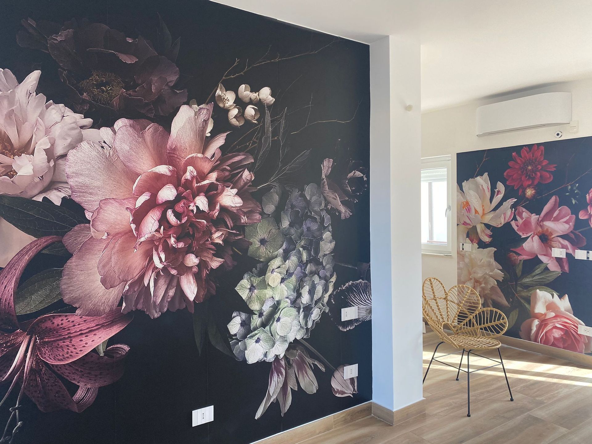 Murale floreale su parete scura, fiori nei toni del rosa, crema e verde. Pareti bianche, pavimento in legno chiaro, sedia dorata.