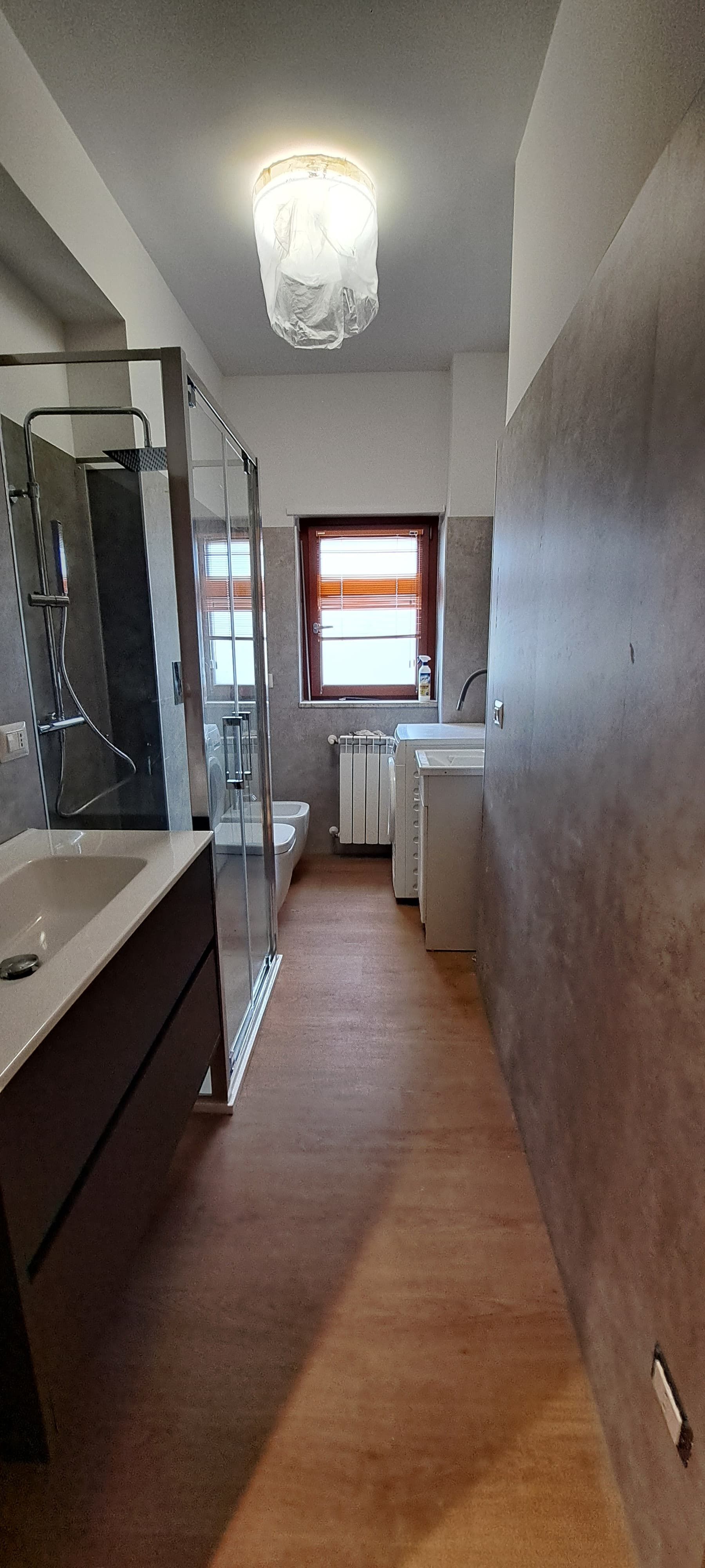 Bagno stretto con doccia, WC e lavandino. Pavimento effetto legno. Lampada decorativa.
