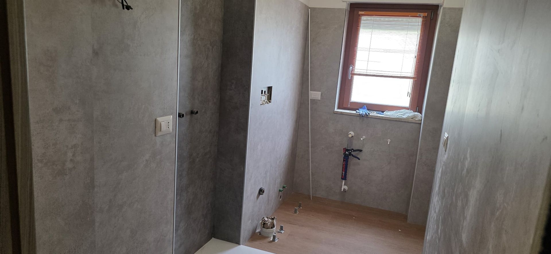 Bagno in costruzione con pareti piastrellate grigie, una finestra e tubature a vista.