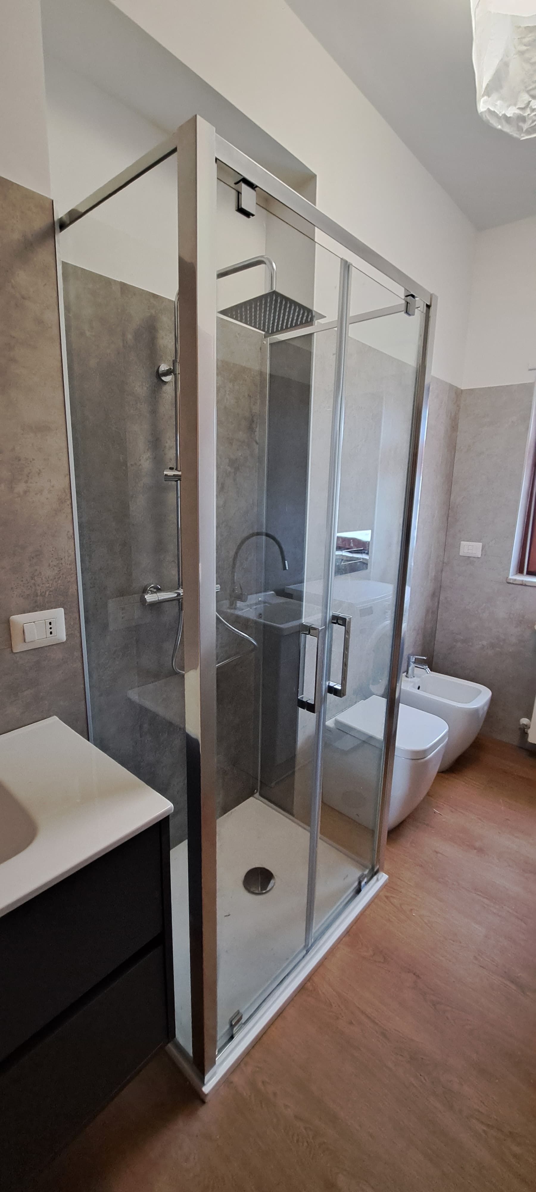 Un bagno moderno con box doccia in vetro, mobile lavabo e bidet. Pavimento in legno. Piastrelle grigie alle pareti.