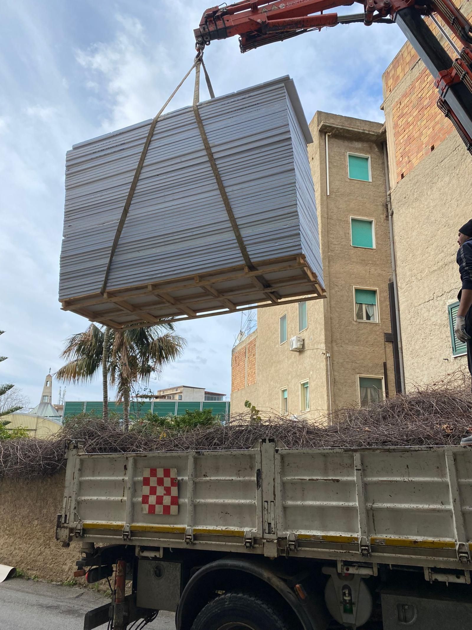 Una gru solleva una grande pila di materiali rettangolari, probabilmente materiali edili, sul cassone di un camion. Edifici sullo sfondo.