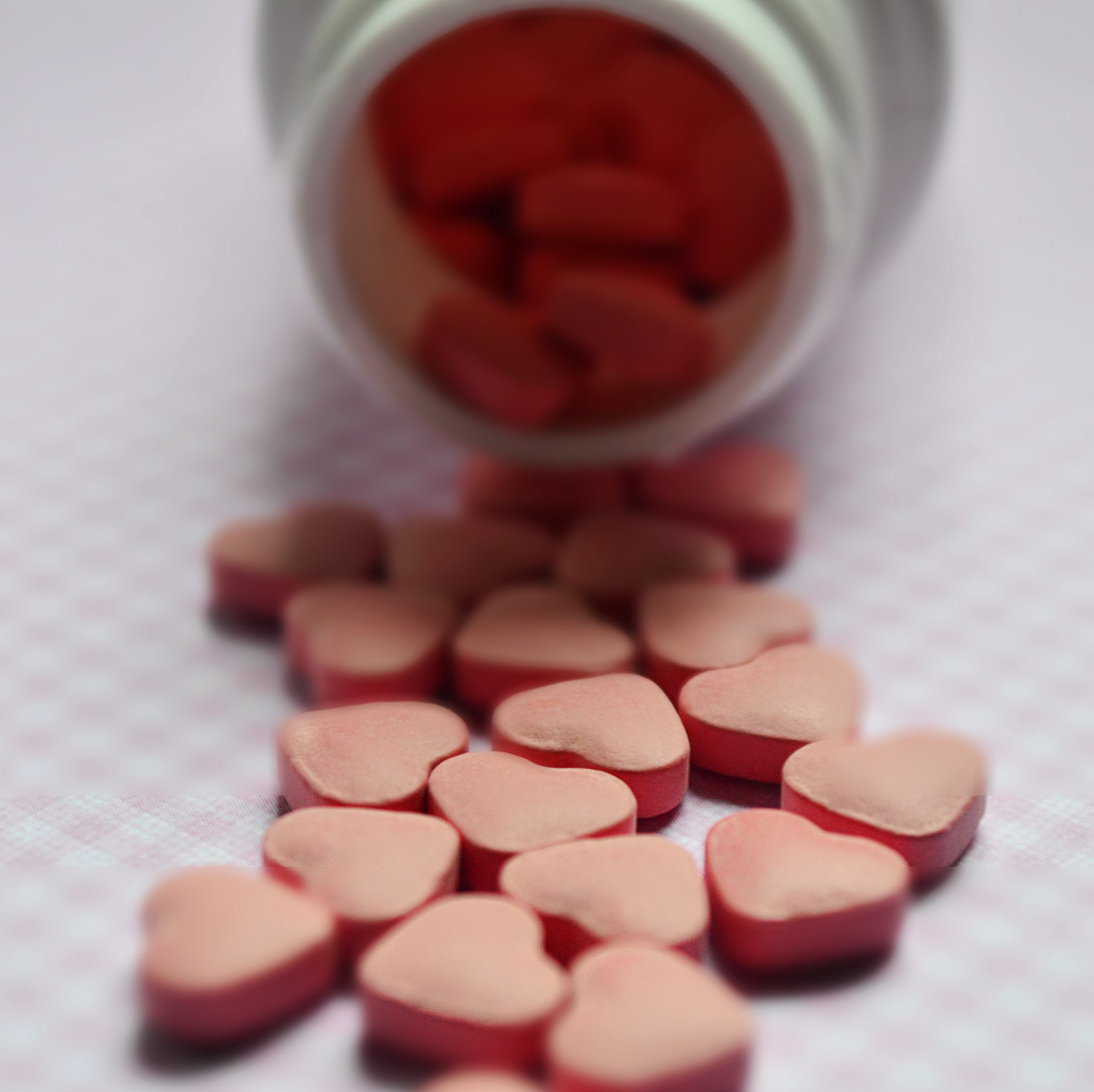 Pink heart-shaped pills spilling from a white bottle onto a pink checkered surface.
