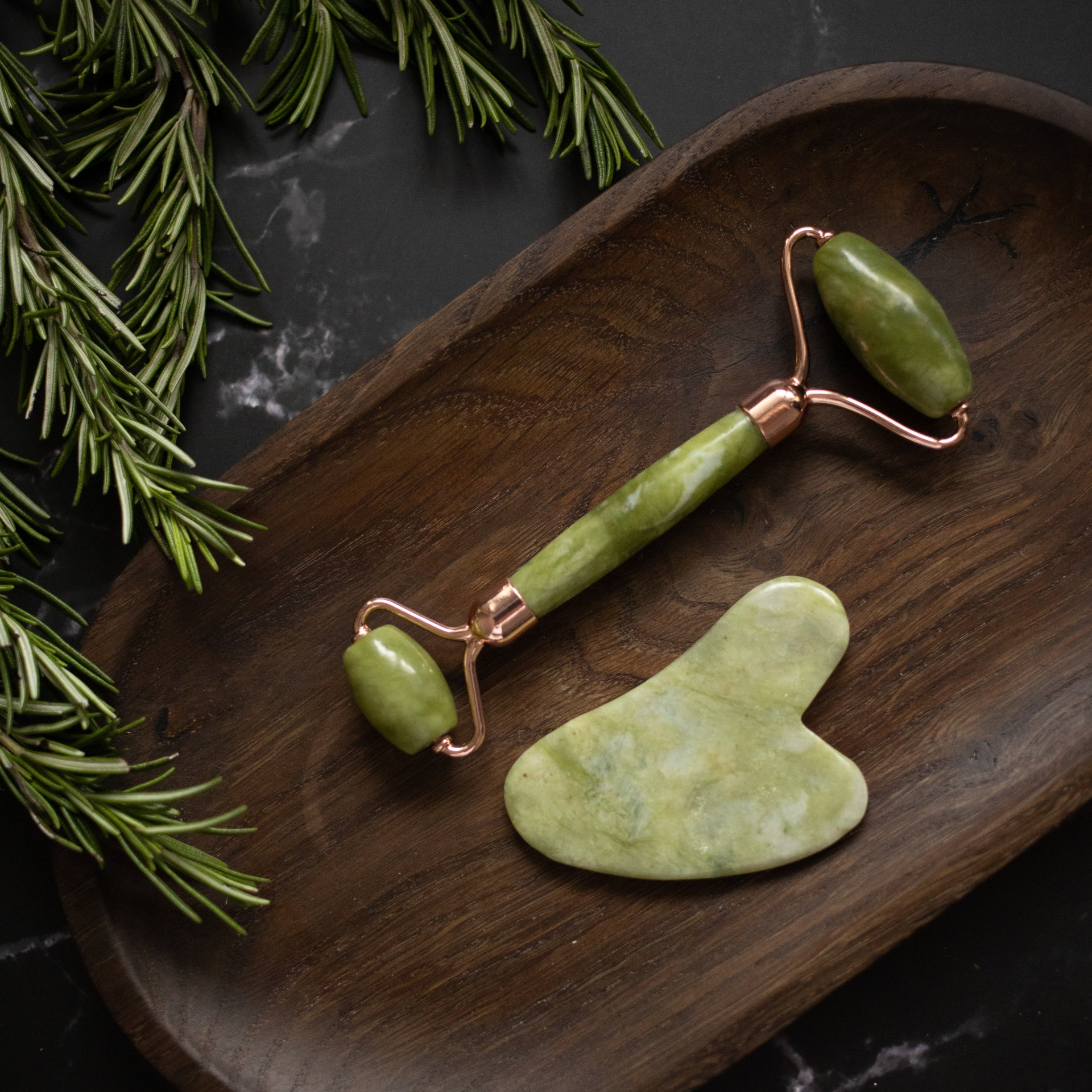 Jade facial roller and gua sha tool on wooden tray with rosemary.