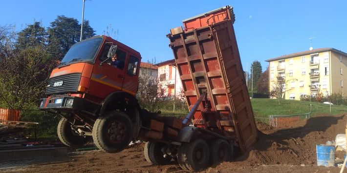 Un camion ribaltabile arancione che scarica il cassone in un cantiere edile sotto un cielo azzurro.