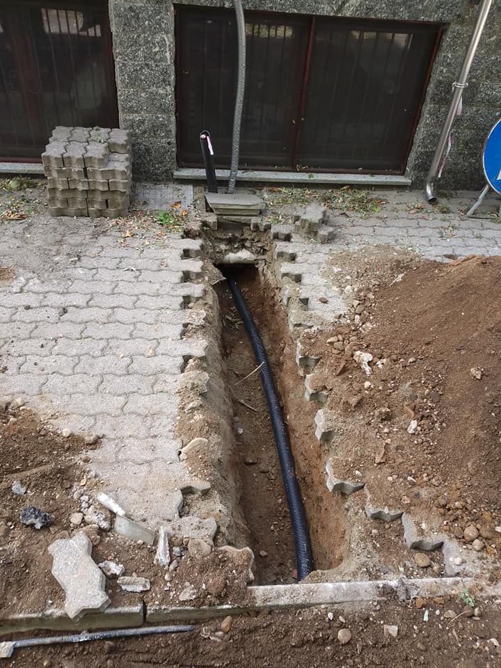 Trincea stretta con tubo nero a vista accanto a un vialetto pavimentato e a un cortile sterrato vicino al muro di un edificio.