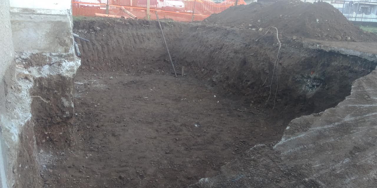 Ampia fossa di scavo con terreno a vista e bordi irregolari, adiacente a un muro di cemento.