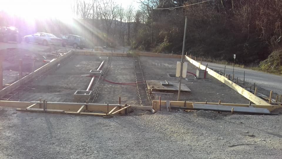 Cantiere edile con ghiaia, fondazioni in cemento e casseforme, esposto alla luce diretta del sole vicino a una strada alberata.