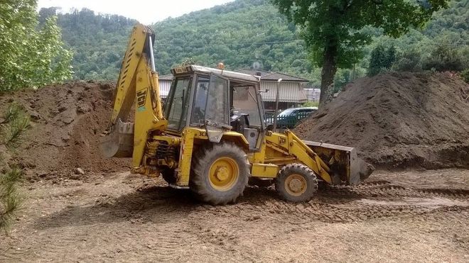 Una terna gialla in un cantiere sterrato con cumuli di terra e alberi sullo sfondo.