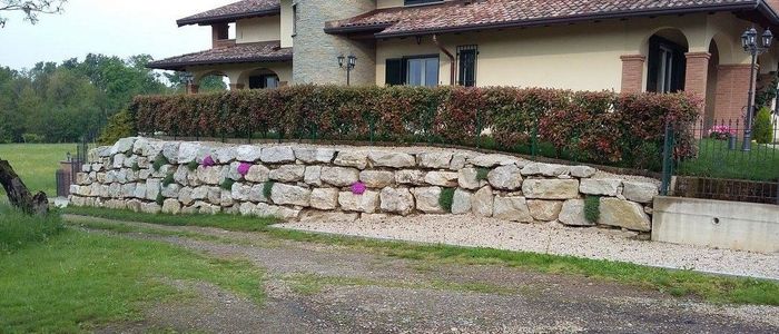 Muro di contenimento in pietra con fiori davanti a una casa rustica e vialetto di ghiaia.