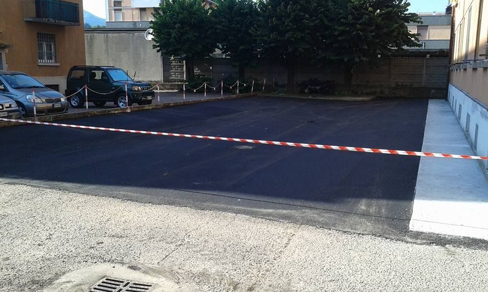 Un cordone di sicurezza delimita un parcheggio vuoto accanto ad alcuni edifici, con auto parcheggiate sullo sfondo.