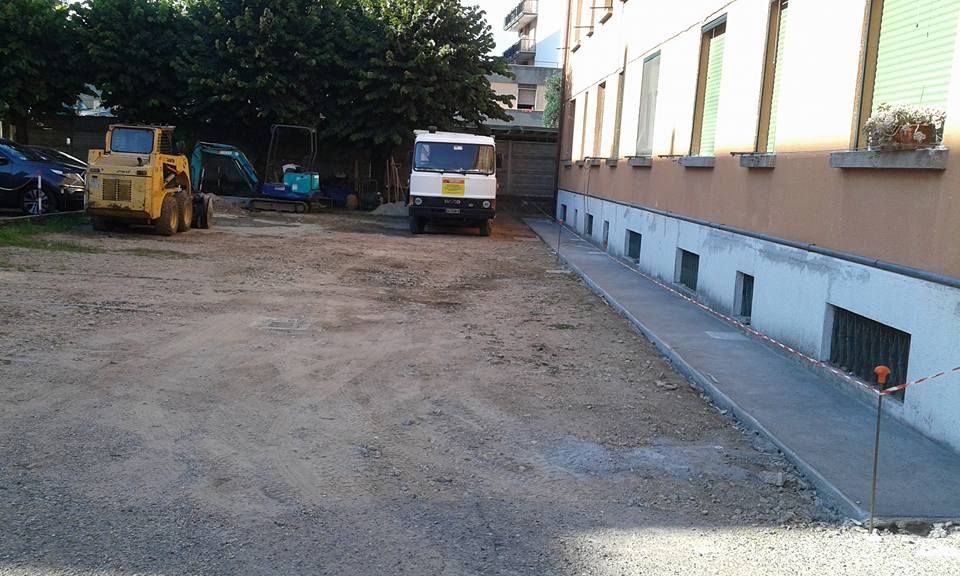 Terreno sterrato accanto a un edificio, con veicoli da cantiere parcheggiati e un furgone bianco vicino al muro.