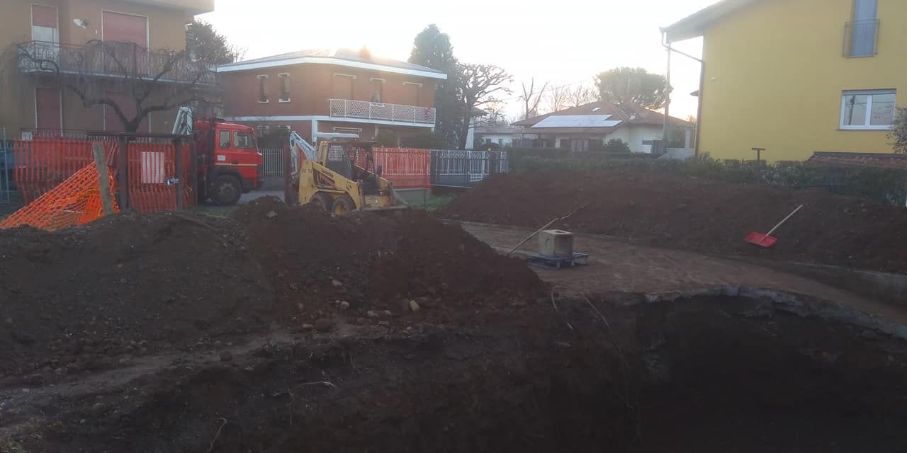Cantiere edile con grandi cumuli di terra, barriere arancioni e case sullo sfondo.