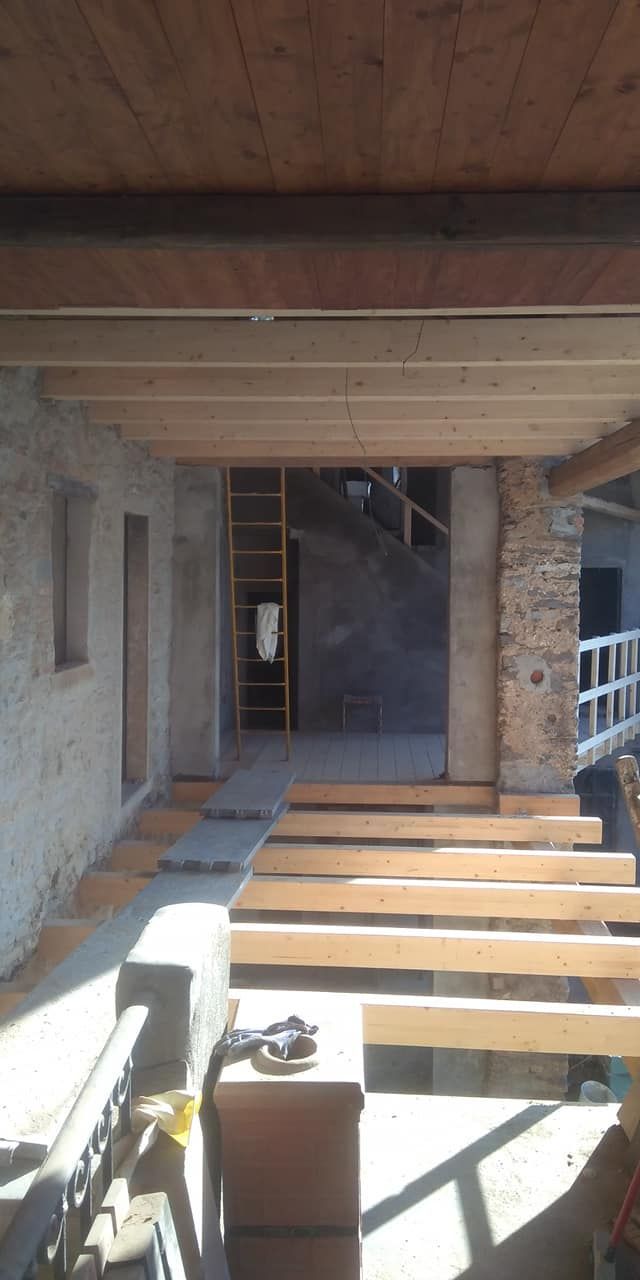 Interno non rifinito con travi del soffitto a vista, muri in pietra e pile di travi di legno sul pavimento.