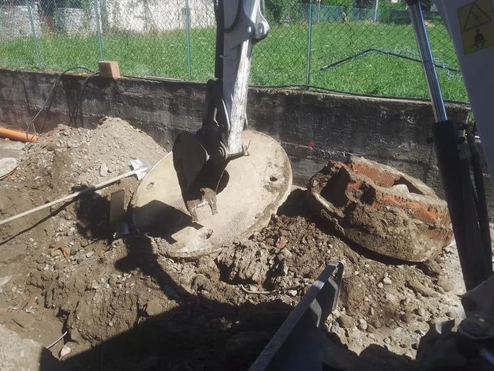 Un escavatore scava terra intorno a un grande oggetto di cemento interrato in un cantiere recintato.
