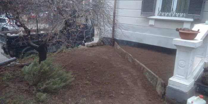Aiuola nel cortile anteriore con terreno nudo, arbusti e ringhiera bianca del portico di una casa.