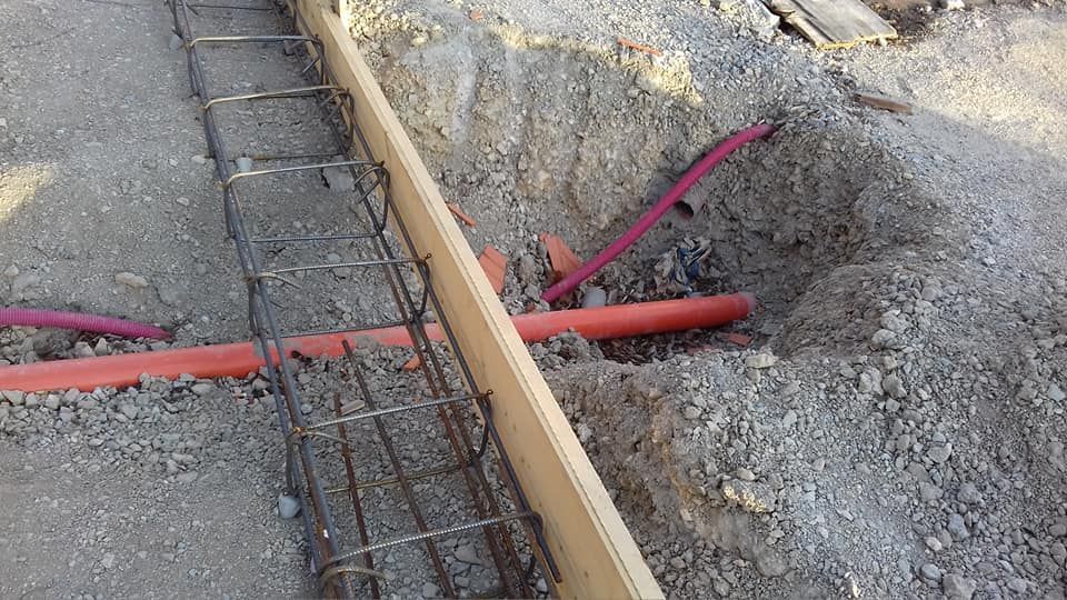 Scavo di costruzione con barre d'armatura, tubi rossi e casseforme per cemento accanto a un marciapiede.