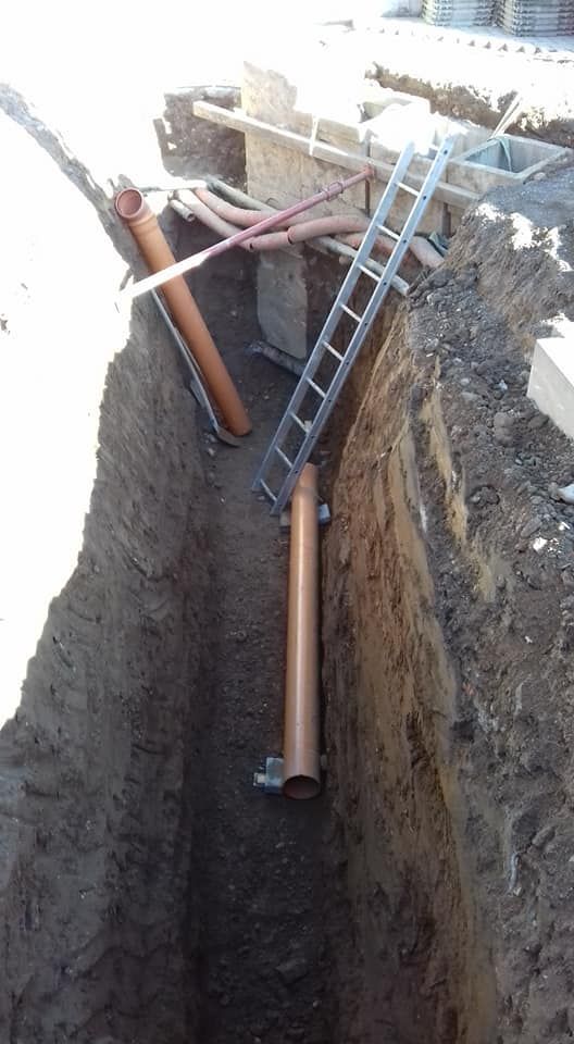 Una profonda trincea con tubi arancioni, una scala e detriti edili in un cantiere.