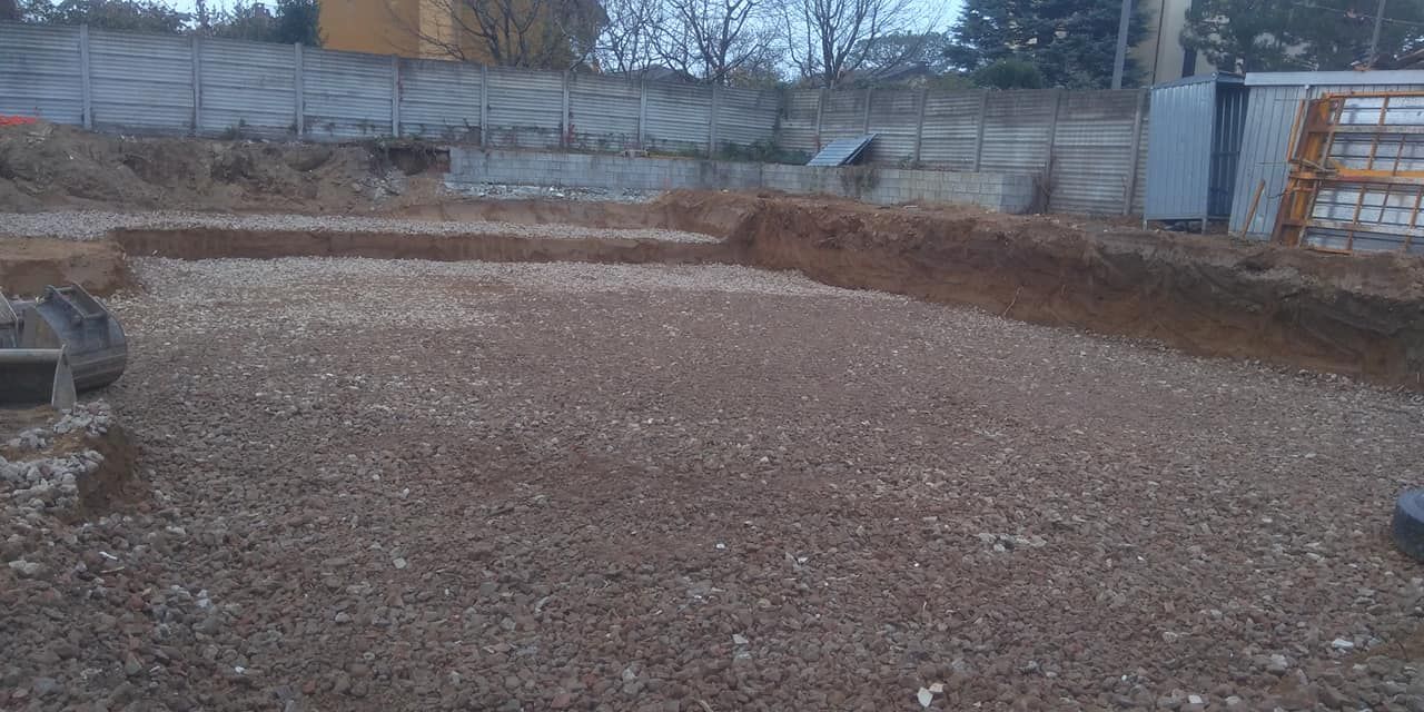 Cantiere edile riempito di ghiaia con bordi di terreno rialzati ed edifici sullo sfondo.
