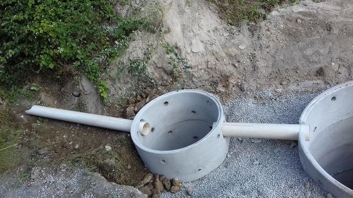 Raccordi per tubi in cemento collegati da un condotto bianco accanto a un terrapieno di terra e ghiaia.