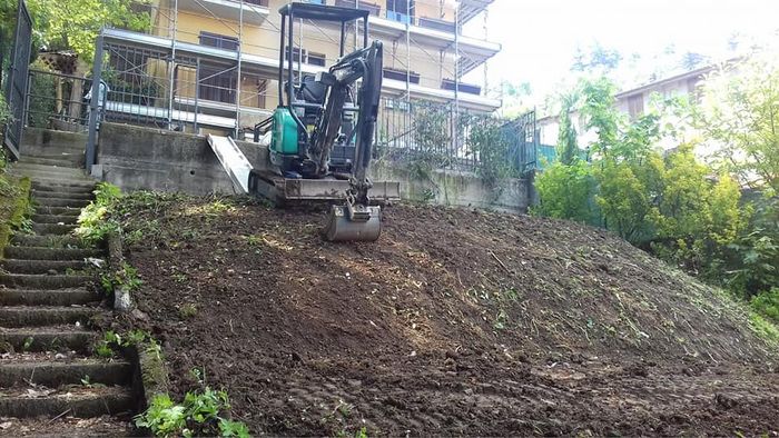 Escavatore su un ripido pendio di terra accanto a scalini di pietra e a un edificio in costruzione.