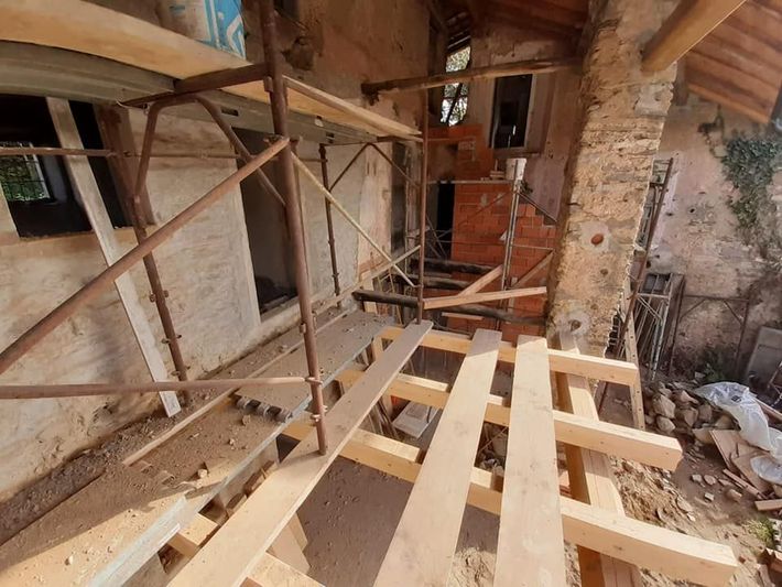 Ponteggi di legno all'interno di un rustico edificio in pietra in fase di ristrutturazione, con travi a vista e detriti sul pavimento.