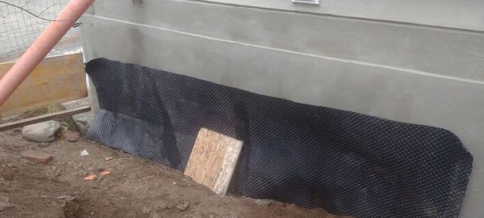 Una rete drenante nera ricopre una trincea di terra accanto a un muro di cemento, con un piccolo paletto di legno a fissarla.