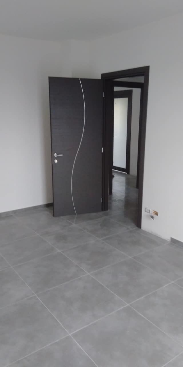 Stanza vuota con pavimento in piastrelle grigie, pareti bianche e una porta scura aperta.