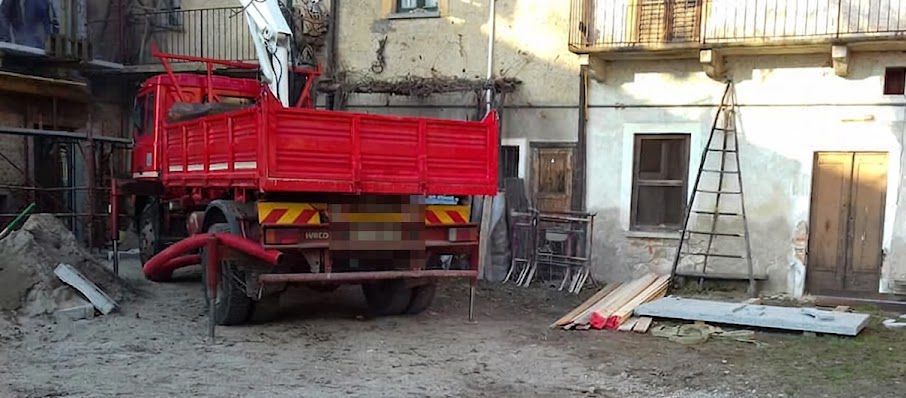 Un camion ribaltabile rosso in un cortile polveroso accanto a vecchi edifici e materiali edili sparsi.