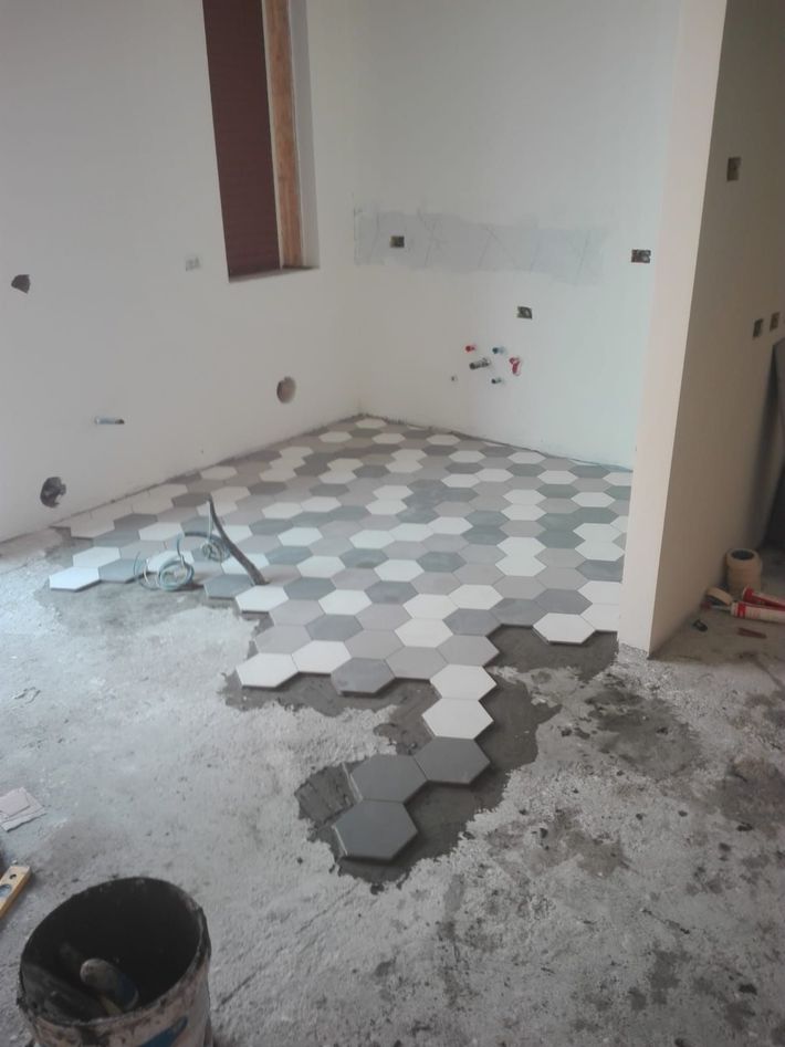 Pavimento della stanza parzialmente piastrellato con piastrelle esagonali grigie in fase di installazione, secchi e attrezzi nelle vicinanze.