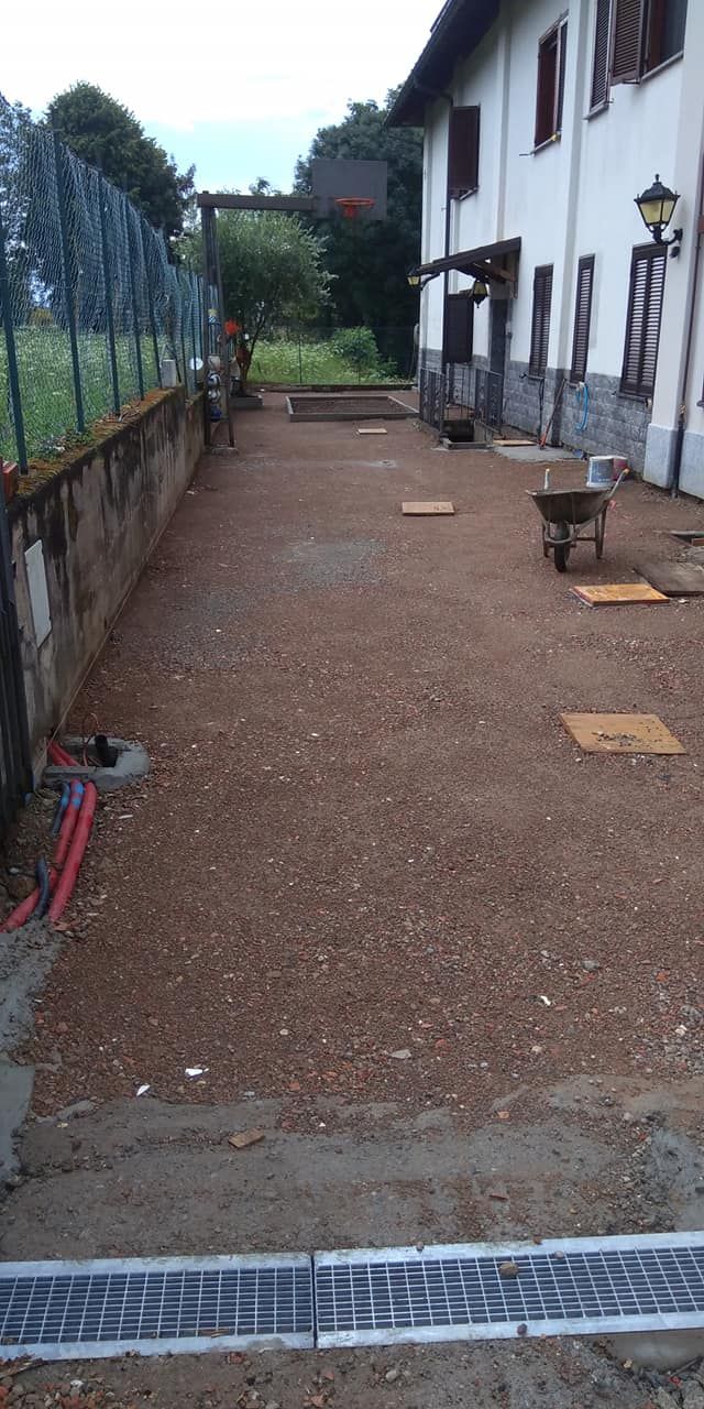 Stretto cortile ghiaioso accanto a un edificio bianco con finestre, una recinzione e una grata di scarico in metallo in primo piano.