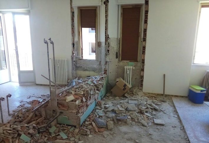 Stanza danneggiata con macerie e sezioni di muro rotte, detriti ammucchiati attorno alla struttura del letto e alle finestre.