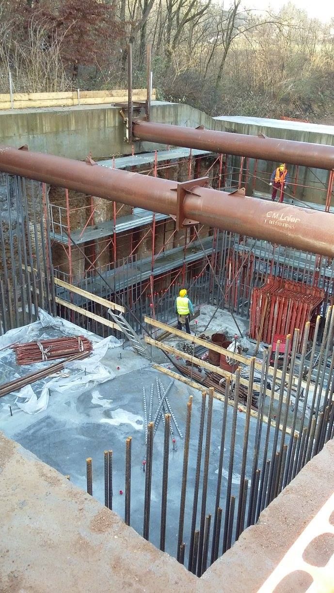 Cantiere edile con scavi profondi, barre d'armatura a vista, supporti in acciaio e un operaio con un giubbotto giallo.
