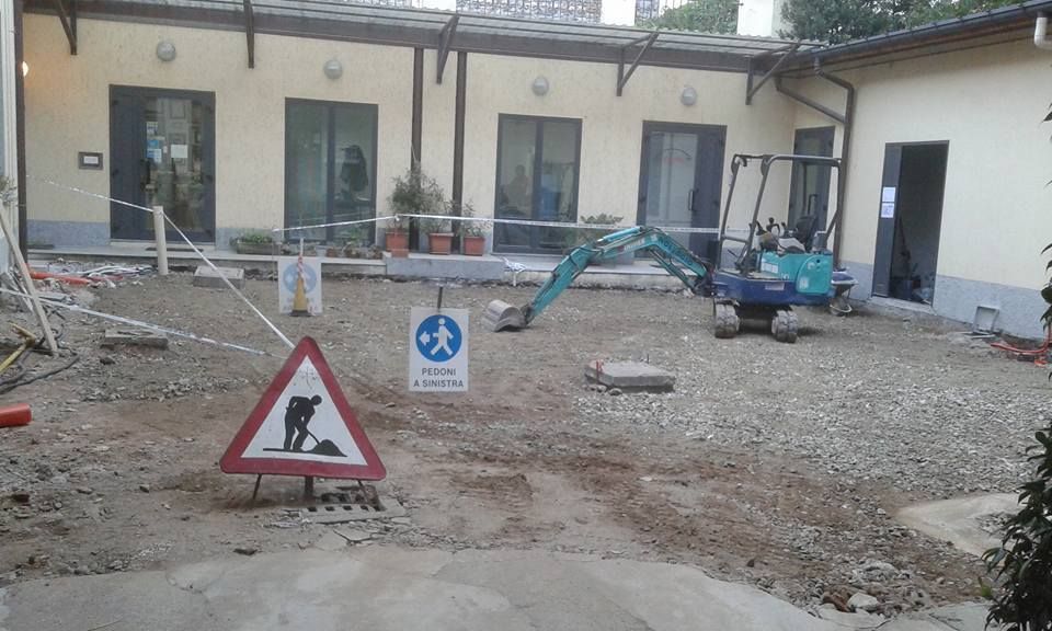 Cantiere edile in un cortile con attrezzi, cartelli e un piccolo escavatore vicino alle porte dell'edificio.