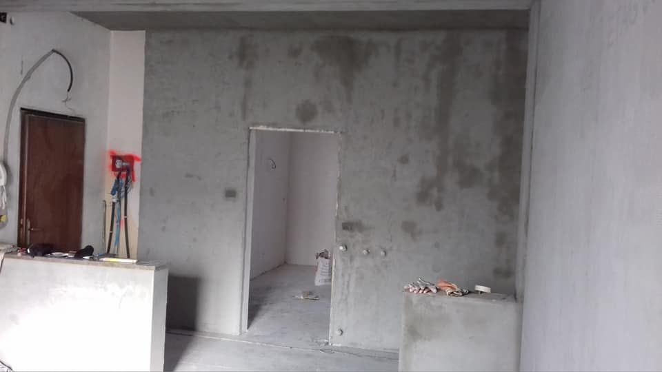 Stanza grigia non rifinita con una porta, pareti in cemento a vista e materiali edili sul pavimento.