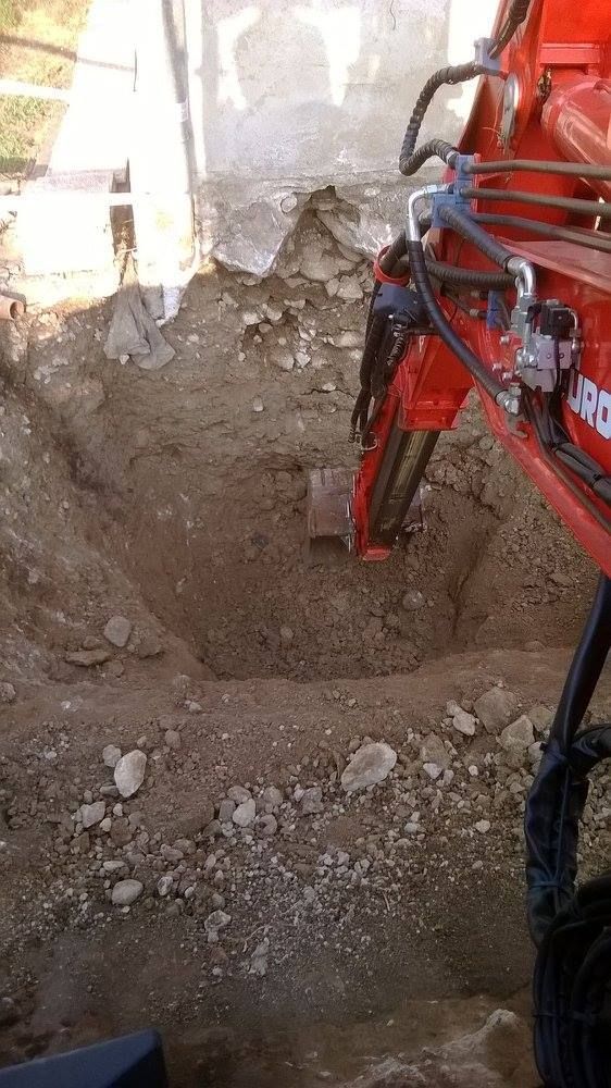 Una ruspa rossa sta scavando una profonda trincea accanto a un muro di terra e detriti.