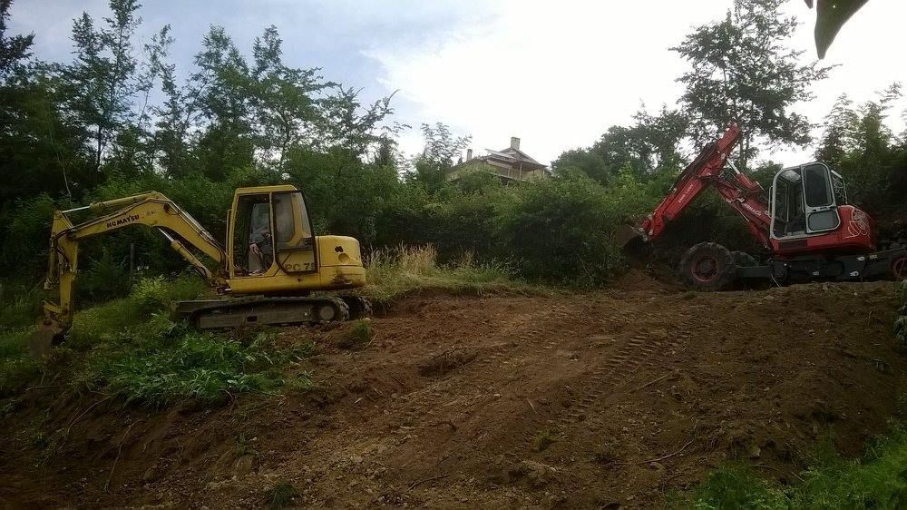 Escavatore giallo e retroescavatore rosso su un pendio di terra in mezzo a una fitta vegetazione verde.
