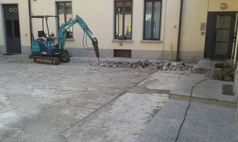 Un mini escavatore sta rimuovendo del cemento in un piccolo cortile accanto a un edificio.