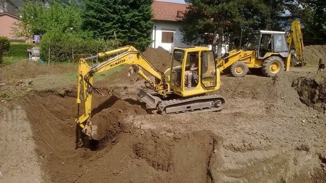 Un escavatore giallo scava una trincea fangosa in un cantiere, con un'altra macchina sullo sfondo.