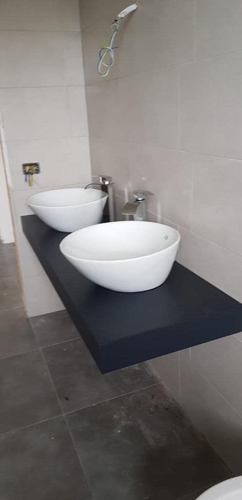 Bagno moderno con due lavabi a ciotola bianchi su un piano di lavoro scuro, a contrasto con le pareti rivestite di piastrelle grigie.