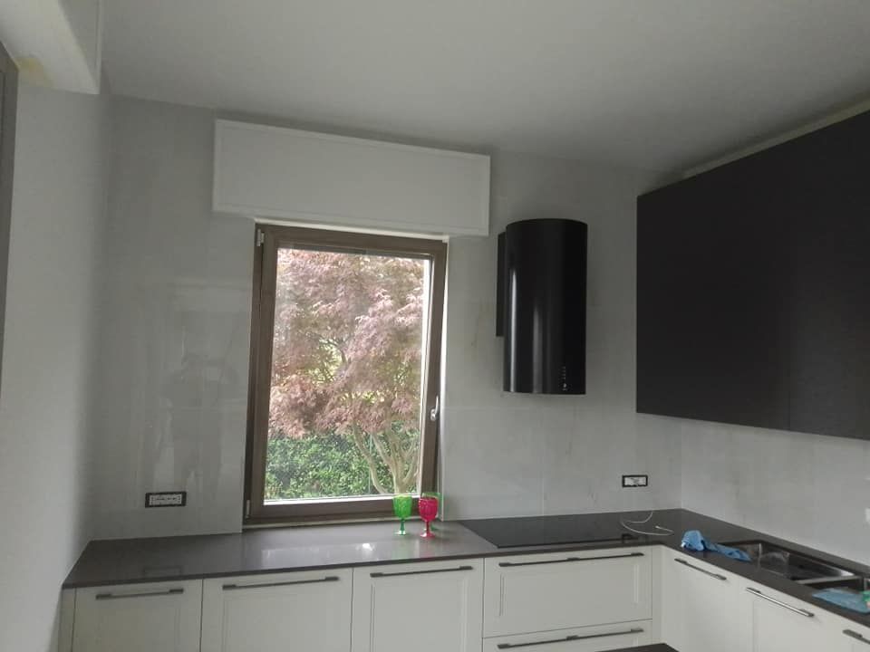 Cucina moderna con mobili bianchi, piano di lavoro grigio, finestra e grande pannello TV nero a parete.