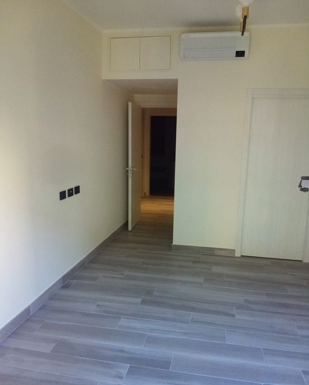Stanza vuota color beige con pavimento in legno grigio, porta aperta e pensili bianchi vicino al soffitto.