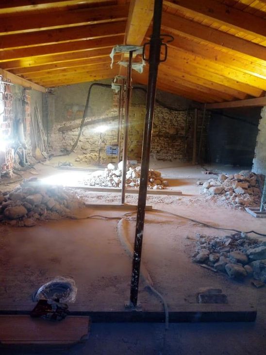 Soffitta abbandonata in fase di ristrutturazione con travi di legno a vista, muri in pietra, detriti e un palo di sostegno centrale.