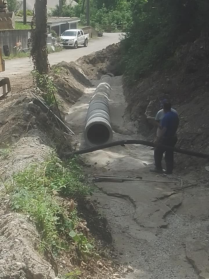 Un operaio maneggia grandi tubi di drenaggio in uno scavo a bordo strada, con un camion nelle vicinanze.