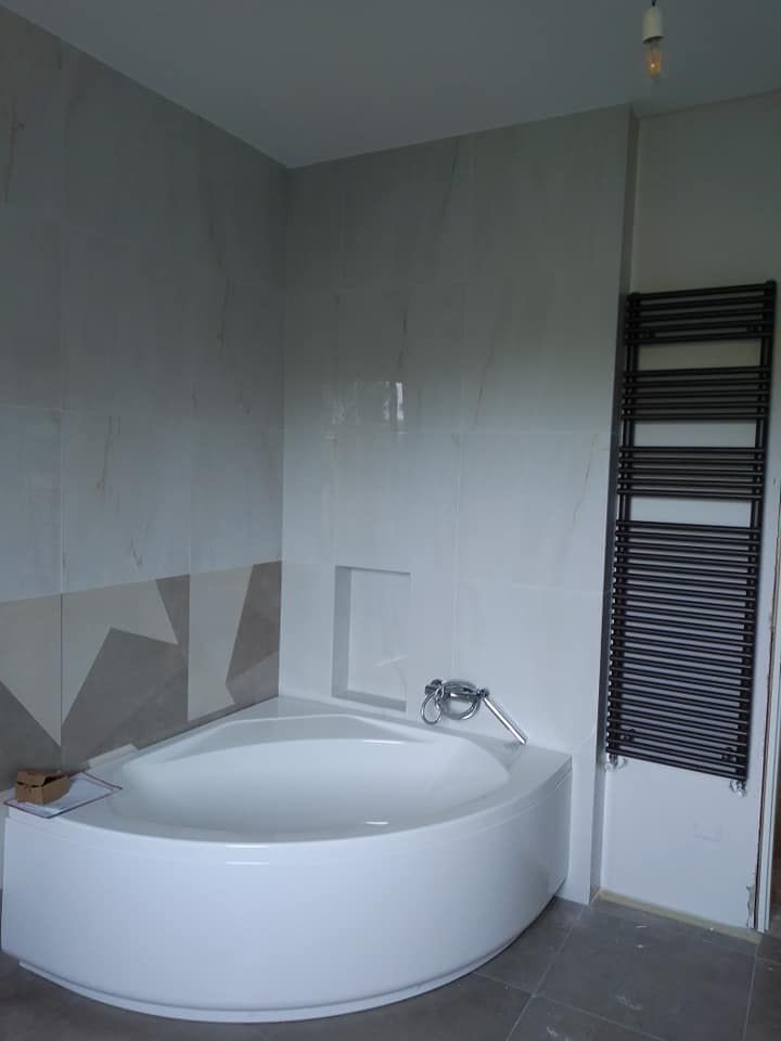 Bagno con una grande vasca angolare bianca, pareti piastrellate e uno scaldasalviette nero.