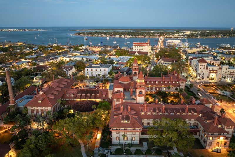 Aerial view of a lit red-roofed coastal town at dusk with a waterfront and harbor beyond.