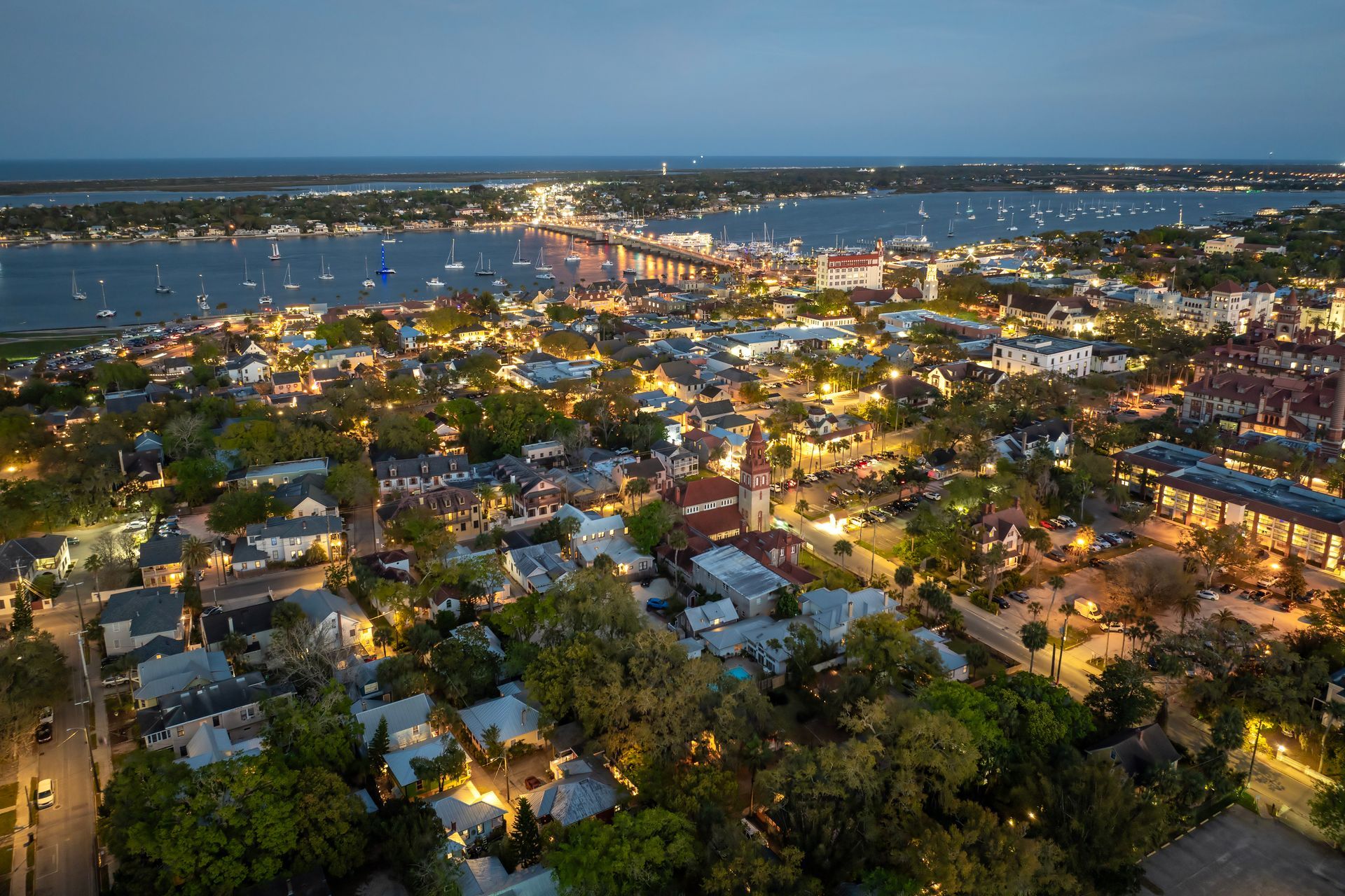 Aerial view of a lit suburban neighborhood at dusk beside a river or bay.